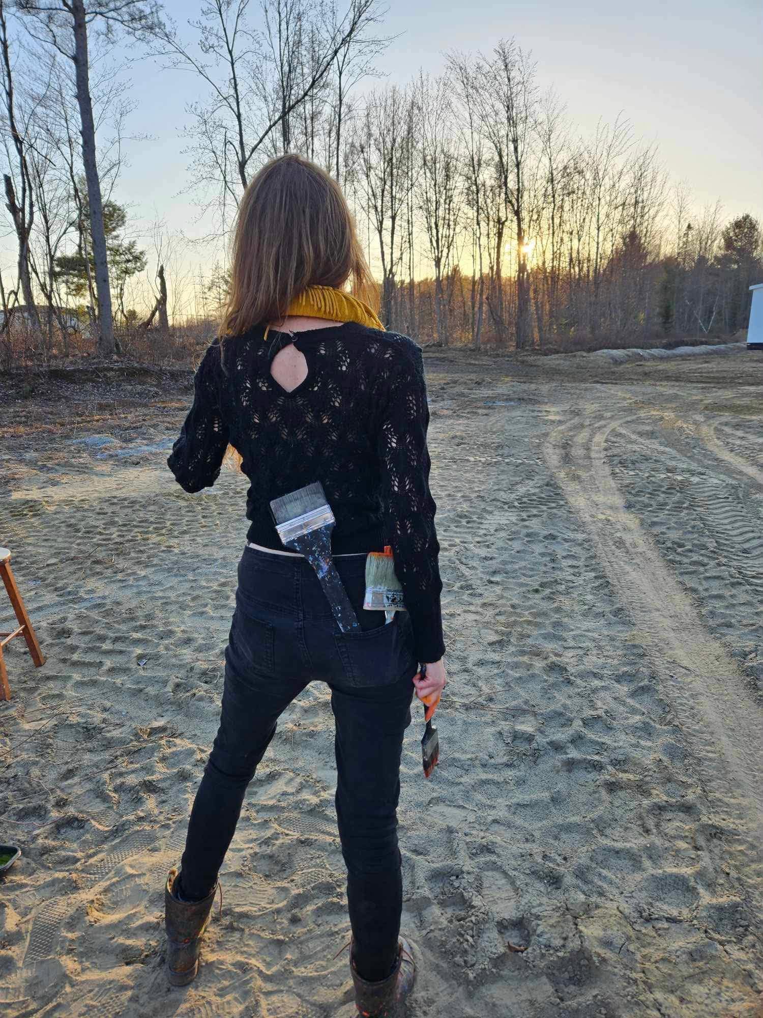 Janet London looking toward a distant sunset in nature, paintbrushes tucked at her back
